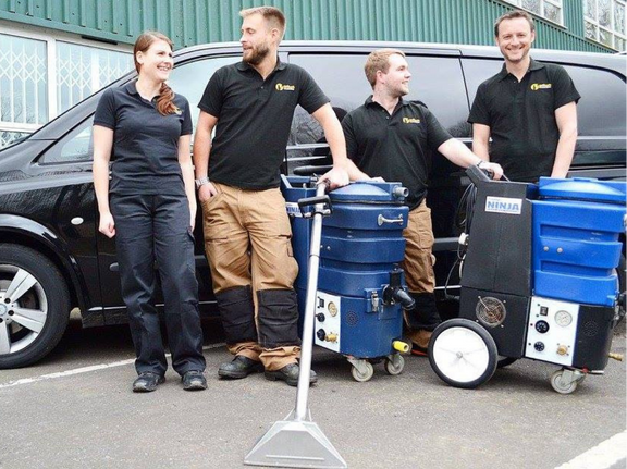 This is a photo of carpet cleaners (three men and one woman) standing in front of their black van, with two steam cleaning carpet machines next to them