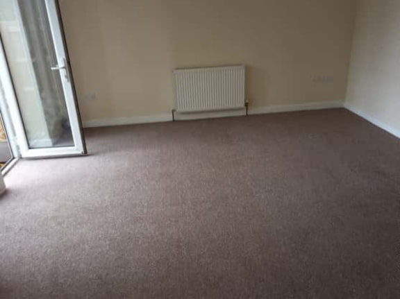 This is a photo of a living room after it has been cleaned. The room is empty and has a brown carpet that has been steam cleaned